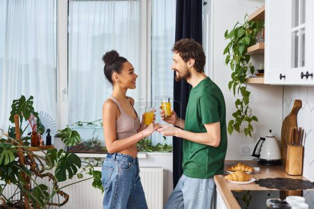 good looking cheerful interracial couple in casual outfits holding glasses of orange juice at home