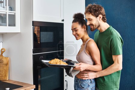 alegre pareja multicultural en ropa de casa utilizando el horno para hornear deliciosos croissants para el desayuno