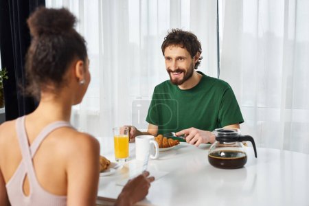 joyous multiracial couple in cozy attires enjoying delicious breakfast together and smiling happily