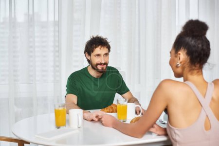 good looking jolly diverse couple in homewear smiling at each other during breakfast at home