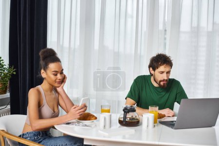 Téléchargez les photos : Séduisante femme afro-américaine regardant le téléphone tandis que son petit ami regardant ordinateur portable au petit déjeuner - en image libre de droit