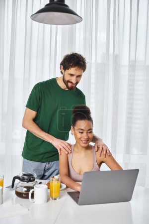 Téléchargez les photos : Séduisant couple multiracial joyeux en utilisant un ordinateur portable pour regarder des films pendant le délicieux petit déjeuner à la maison - en image libre de droit