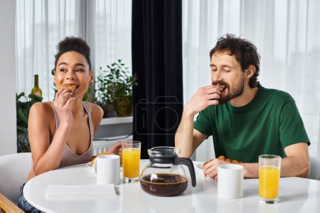 Foto de Hermosa alegre pareja diversa en ropa de casa disfrutando de su delicioso desayuno mientras está en casa - Imagen libre de derechos