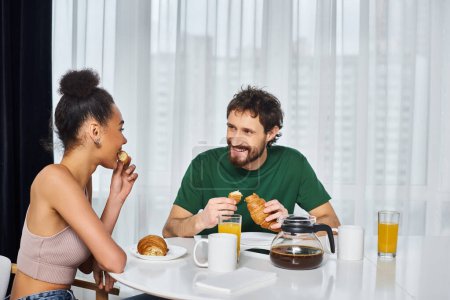 Téléchargez les photos : Attrayant couple joyeux multiculturel en vêtements de maison ayant délicieux petit déjeuner ensemble à la maison - en image libre de droit