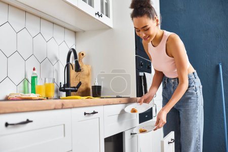 bien parecido alegre africana americana mujer en la limpieza de la ropa de casa platos después del desayuno en casa