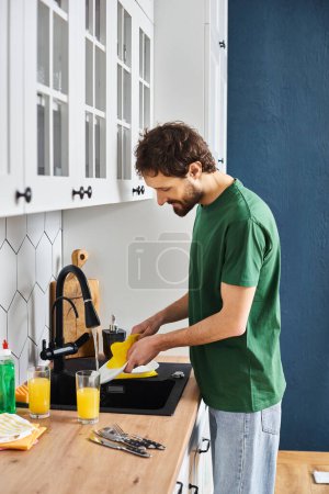 cheerful handsome man in comfortable homewear washing plates after breakfast at home in kitchen