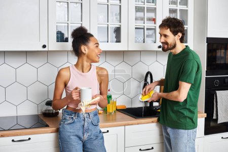 alegre multicultural hermosa pareja en la ropa de casa lavar los platos y sonriendo el uno al otro en casa
