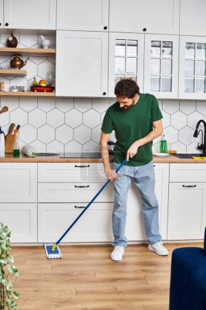 Foto de Hombre alegre guapo en traje casual cómodo usando fregona para limpiar el piso mientras está en la cocina en casa - Imagen libre de derechos