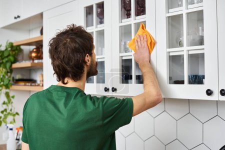 back view of man in casual dark green t shirt using rag to clean kitchen cupboards while at home
