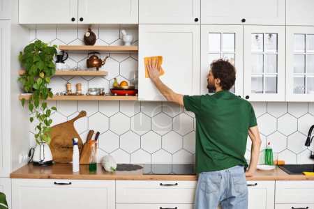 Foto de Vista trasera del hombre en cómoda camiseta verde con trapo amarillo para limpiar los armarios de la cocina en casa - Imagen libre de derechos