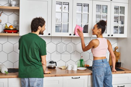 séduisant interracial couple joyeux dans des tenues décontractées laver les surfaces dans la cuisine, nettoyage de printemps