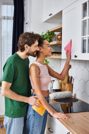 beautiful jolly diverse couple in cozy outfits doing chores in kitchen together and smiling happily