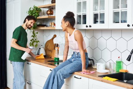 cheerful african american woman sitting on counter and looking at her boyfriend cleaning kitchen