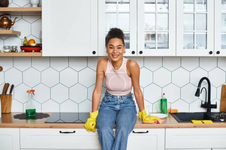cheerful african american woman in homewear sitting on kitchen counter and looking at camera