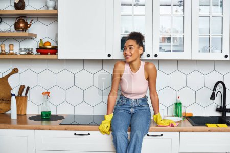 joyeuse femme afro-américaine attrayante en vêtements de maison assis sur le comptoir de la cuisine et regardant loin