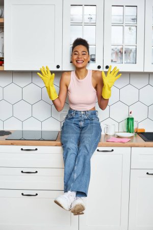 alluring jolly african american woman in homewear sitting on kitchen counter and smiling at camera