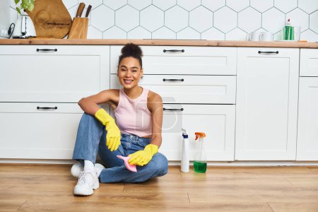 cheerful good looking african american woman in casual outfit sitting on floor and smiling at camera
