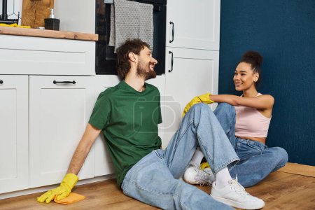 joyful diverse beautiful couple in homewear sitting on floor and smiling happily on spring cleaning