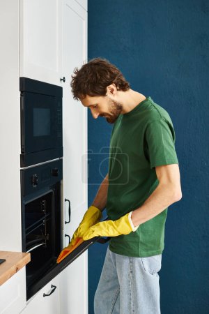 handsome cheerful man in dark green t shirt and gloves cleaning modern oven with yellow rag