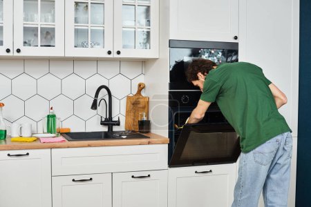 Foto de Hombre atractivo en traje acogedor diario con guantes usando trapo para lavar el horno durante la limpieza de primavera - Imagen libre de derechos