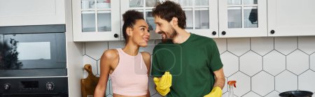 jolly diverse couple in comfortable homewear smiling at each other during spring cleaning, banner