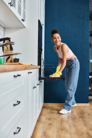 cheerful beautiful african american woman in casual outfit washing oven in kitchen, spring cleaning