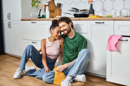 interracial jolly couple in casual attires sitting on floor during cleaning and smiling happily
