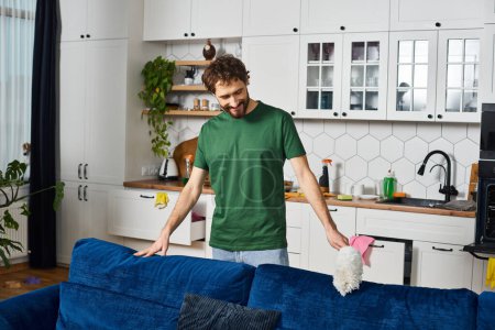 Foto de Hombre guapo alegre en acogedor sofá de limpieza de ropa de casa con plumero y sonriendo felizmente mientras está en casa - Imagen libre de derechos