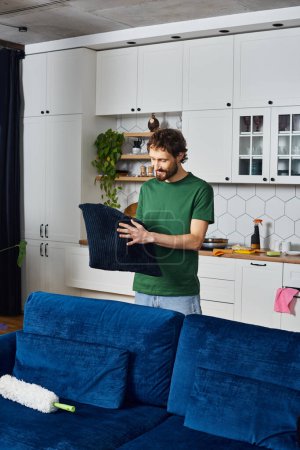 Foto de Atractivo hombre alegre en camiseta verde casual sosteniendo almohada en sus manos mientras limpia en casa - Imagen libre de derechos