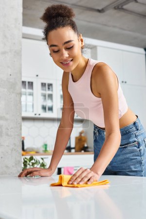 cheerful beautiful african american woman in cozy homewear using rag to clean table at home