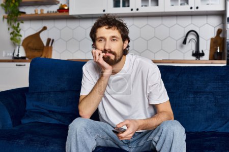 Foto de Hombre guapo aburrido en acogedora ropa de casa sentado en el sofá con control remoto en la mano y viendo la televisión - Imagen libre de derechos