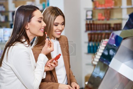 joyful attractive women in business casual attires choosing new lipstick in cosmetics store