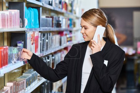jolly blonde beautiful saleswoman in black blazer smiling and talking by smartphone in store