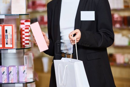 cropped view of elegant saleswoman in black stylish blazer putting new perfumes into shopping bag