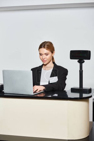Foto de Atractiva vendedora con estilo en blazer negro trabajando en el ordenador portátil mientras está sentado en el mostrador en la tienda - Imagen libre de derechos