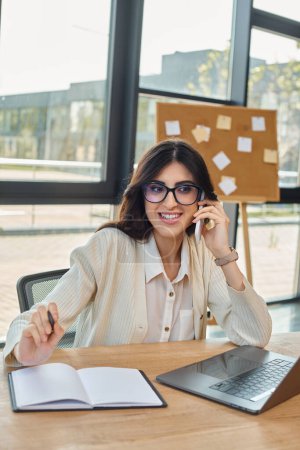 Photo for A professional businesswoman, part of the franchise concept, sitting at a desk in a modern office, talking on her cell phone. - Royalty Free Image