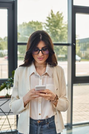 A focused businesswoman stands by a window, checking her cell phone in a modern office setting.