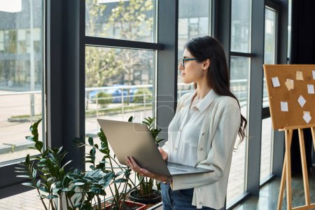 A modern businesswoman stands by a window, holding a laptop in her hands as she works on franchise concepts.