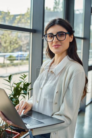 Photo for A sophisticated woman in glasses confidently holds a laptop in a modern office, embodying innovation in the business world. - Royalty Free Image