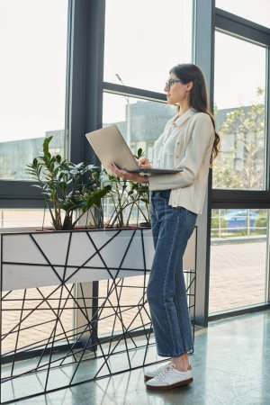 A businesswoman stands by a window, focused on her laptop in a modern office space with a franchise concept.