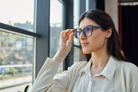 A businesswoman, wearing glasses, gazes out a window in a modern office, contemplating the urban landscape.