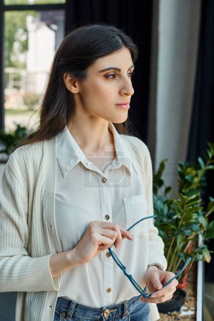 Una mujer de negocios se para con confianza frente a una ventana, mirando hacia el exterior en un moderno entorno de oficina cerca de su espacio de trabajo.