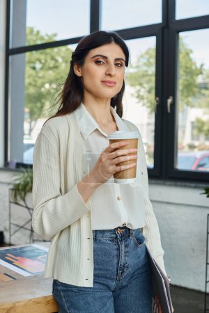 A modern businesswoman stands by a window, holding a cup of coffee.