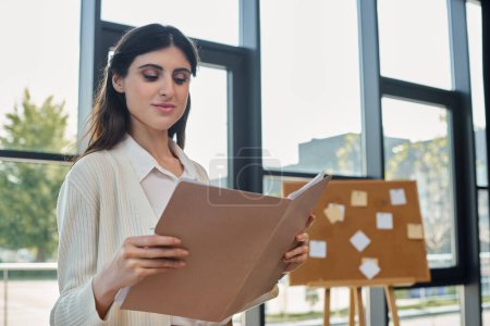 A businesswoman stands near her workspace, holding a paper in front of a window, contemplating her next steps in the franchise world.