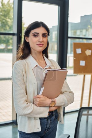 A businesswoman standing confidently in a modern office, holding a binder next to a table.