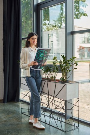 A modern businesswoman stands near her workspace, holding charts, gazing out of a window.