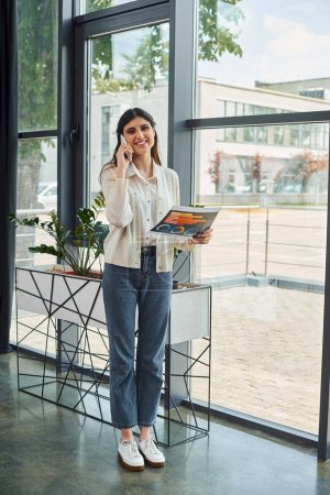 Eine Geschäftsfrau steht in einem modernen Büro am Fenster, telefoniert und präsentiert ein Franchise-Konzept.