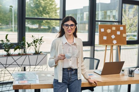 Téléchargez les photos : Une femme d'affaires se tient dans un bureau moderne, concentré sur son ordinateur portable sur la table devant elle. - en image libre de droit