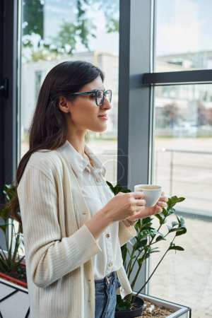 A modern businesswoman holds a cup, standing in front of a window in an office, deep in thought.