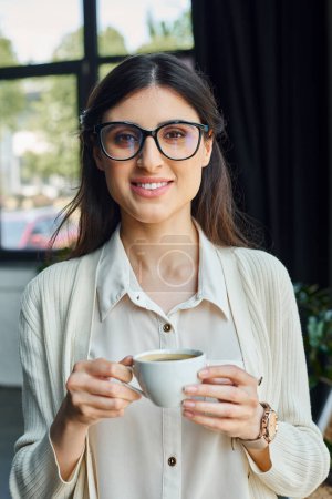 A businesswoman with glasses enjoys a coffee break in a modern office workspace.